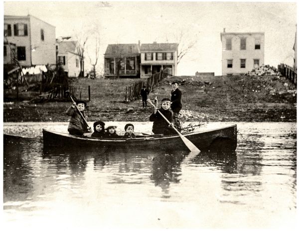 Image of five children rowing a canoe, with houses in  the background.