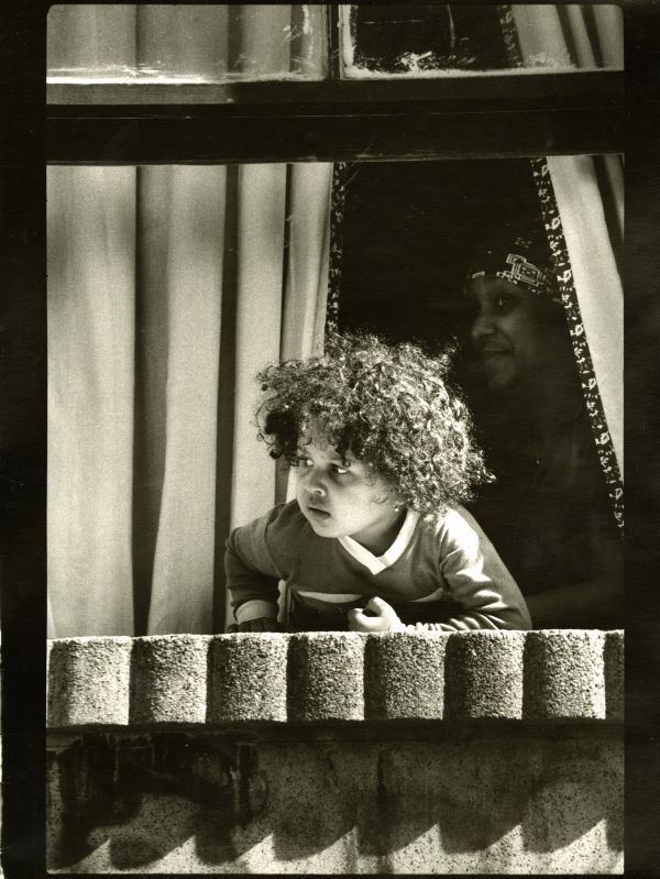 Image of a child looking out the window to his left while his mother, partially obscured by shadow, looks at the camera.