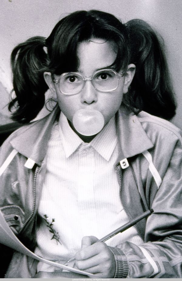 Image of girl blowing a bubble with bubble gum. She wears glasses and pigtails.