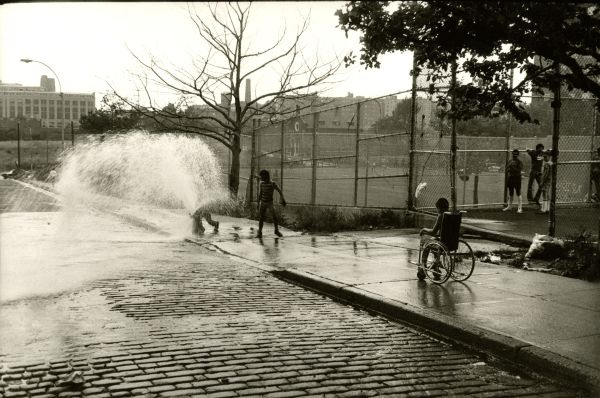 Image of children, including one on a wheelchair, gathered near an opened fire  hydrant.