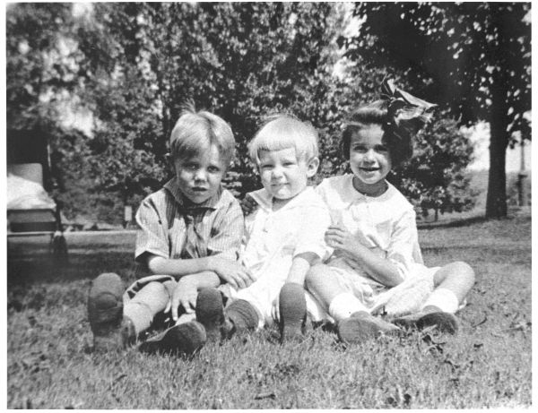 Image of three children sitting on the grass and looking at the camera. A baby carriage is behind them.
