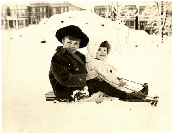 Image of two children sledding on Grand Concourse. There are houses behind them.
