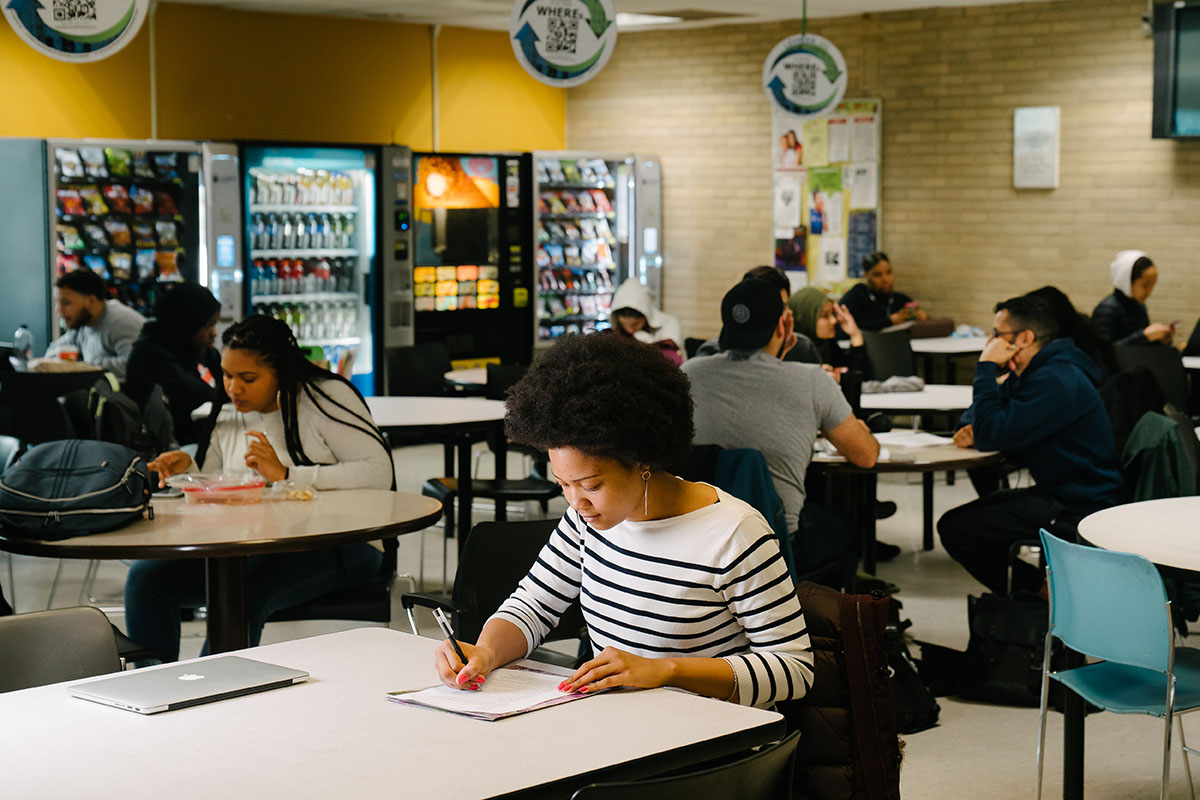 A student studying and writing in Carman Cafeteria, surrounded by peers and vending machines.