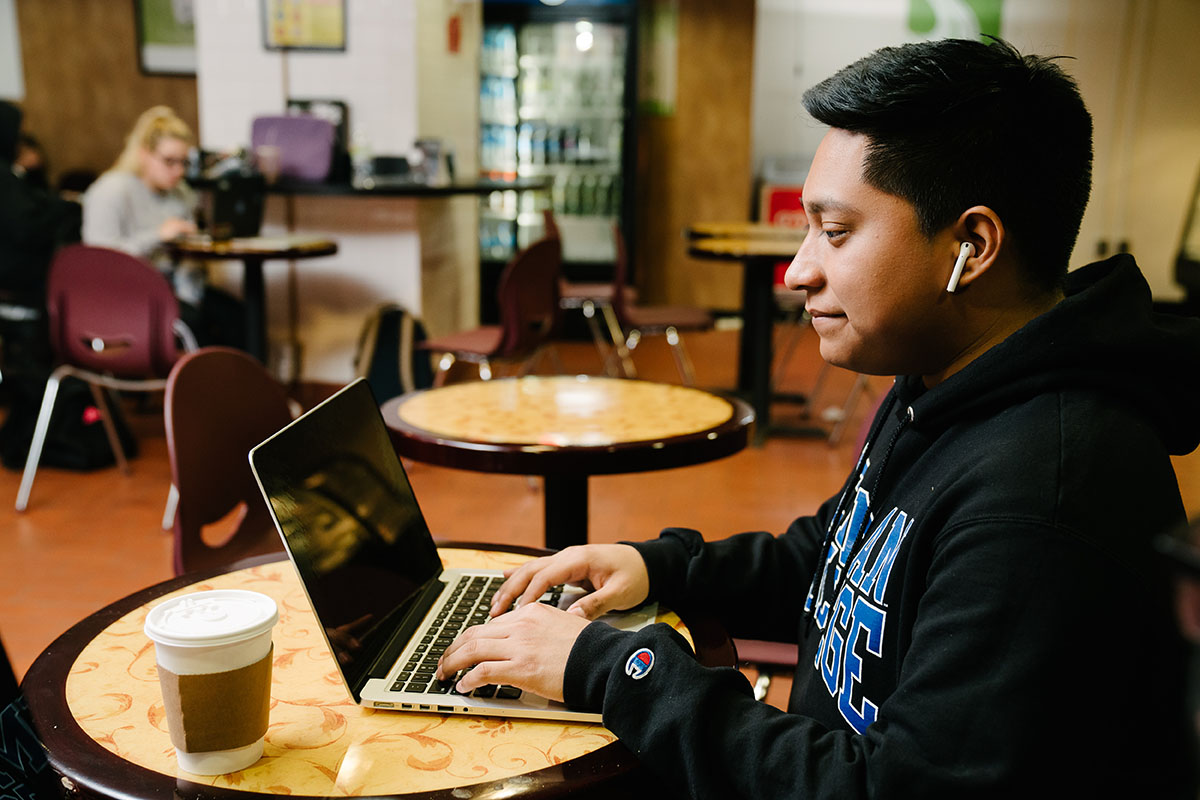 Lehman College student wearing a black hoodie and wireless earbuds works on a laptop at Harmony Café table.