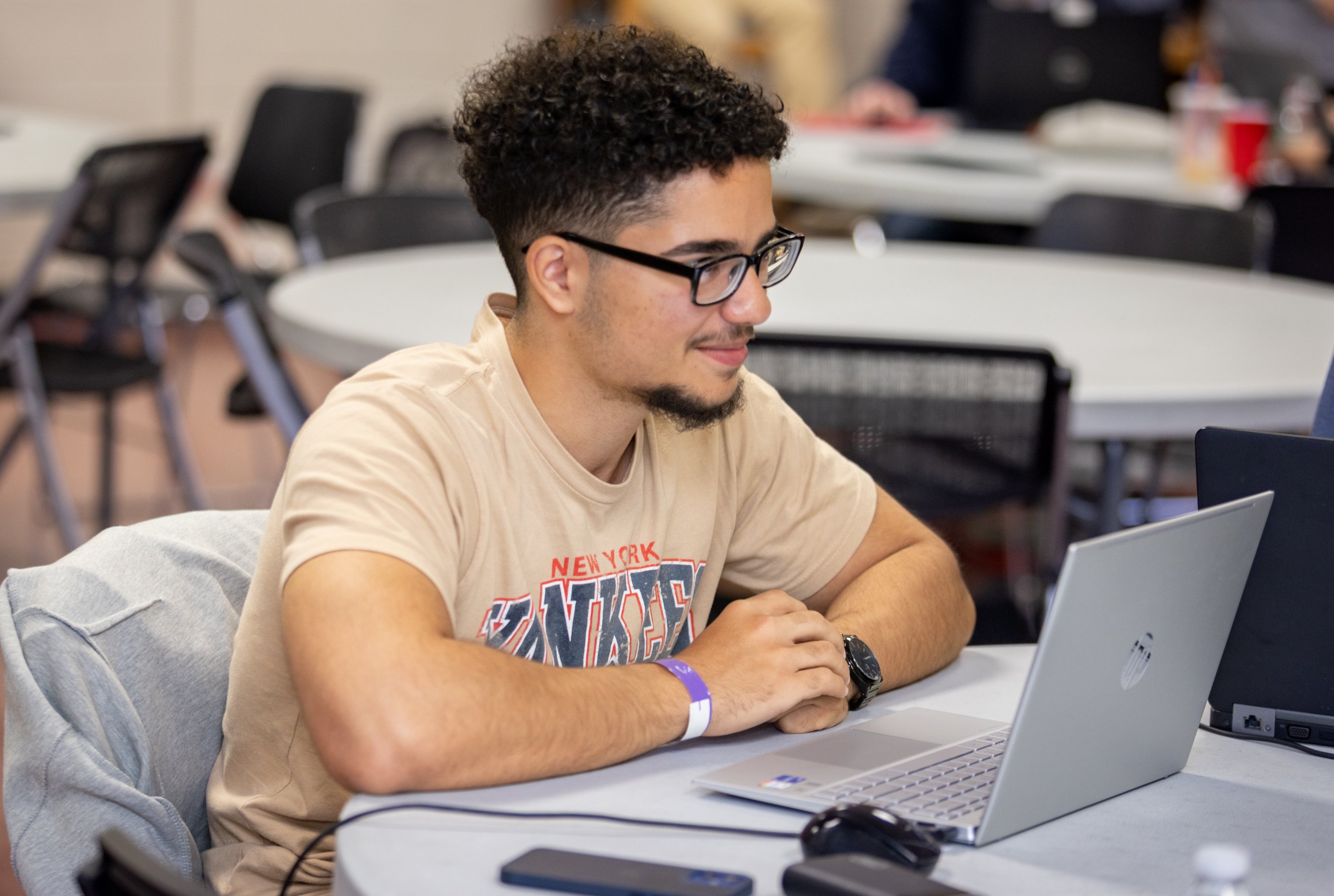 A student wearing glasses smiling while working on a laptop at a table during Hackathon event.