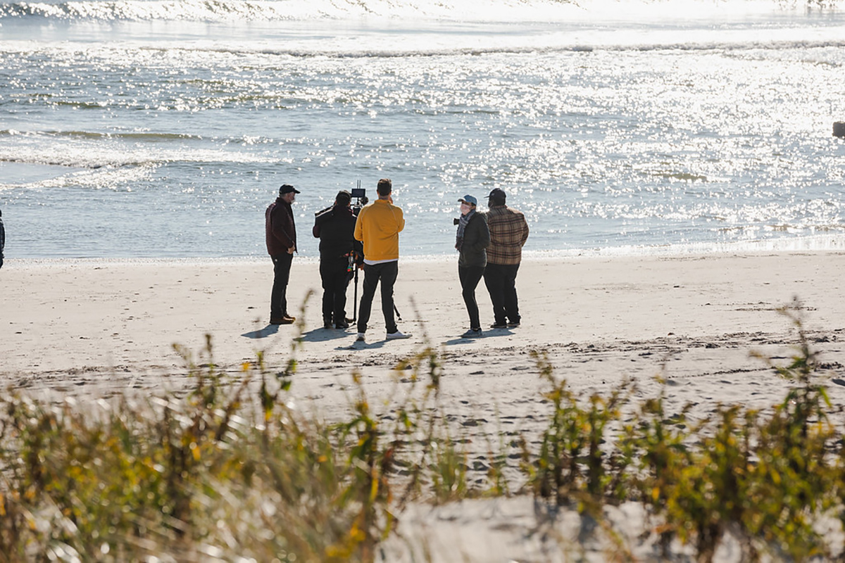 A film crew standing on a sandy beach near the ocean shoreline filming on a sunny day.