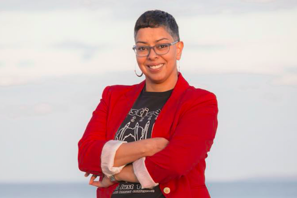 Woman with short hair and glasses in red jacket standing with arms folded.