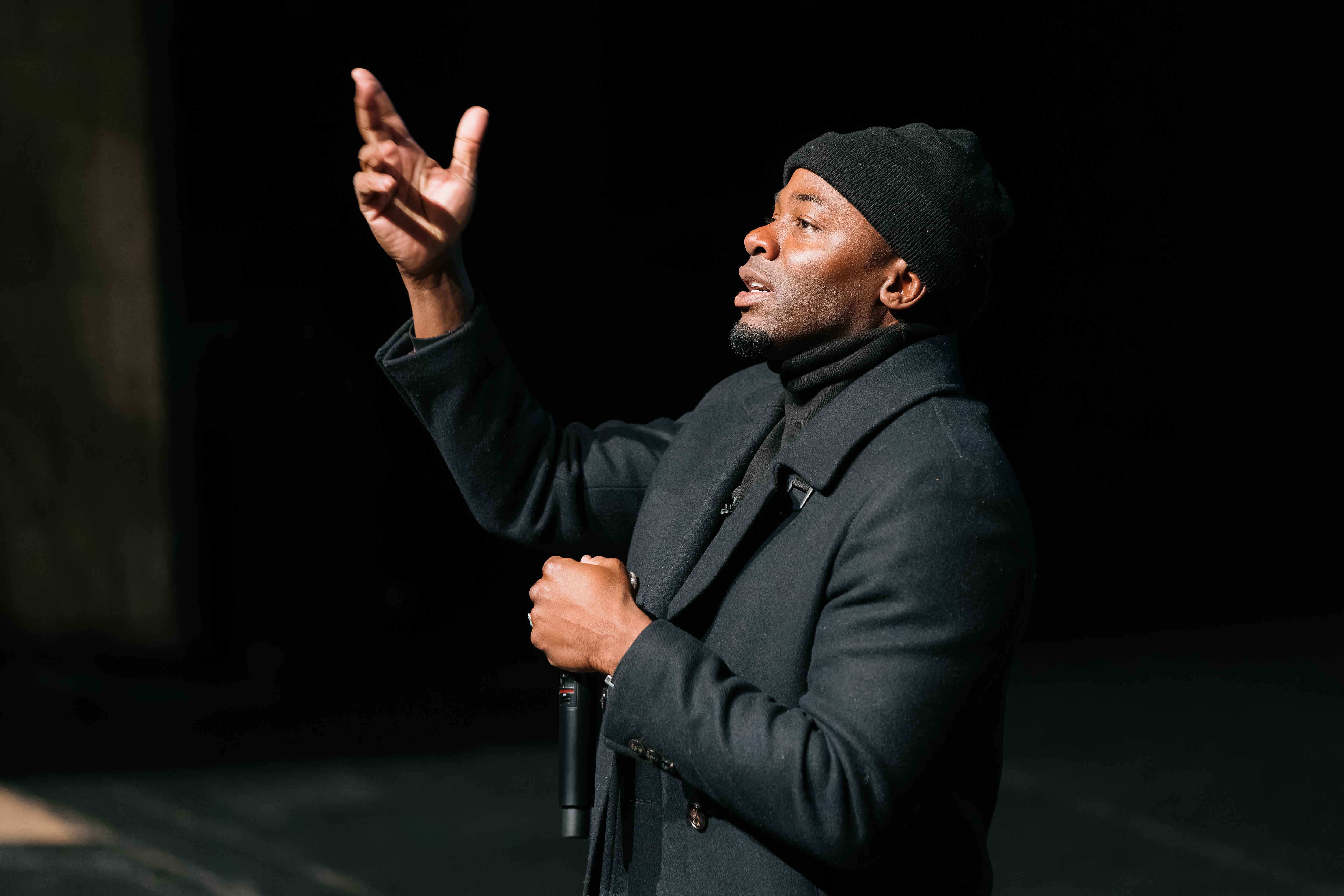 Photo of an African American man in profile. He is dressed in black and gesturing in the air.