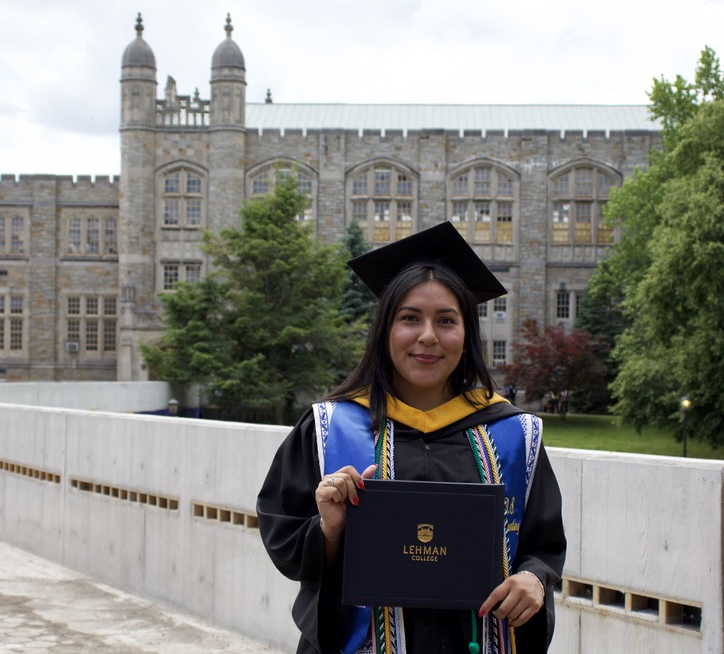 Maria Campos in commencement regalia holds her diploma with Lehman College's Old Gym in the background.