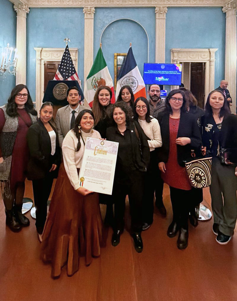 A group of people posing with a mayoral citation. U.S., Mexican, and New York State flags are displayed behind them.