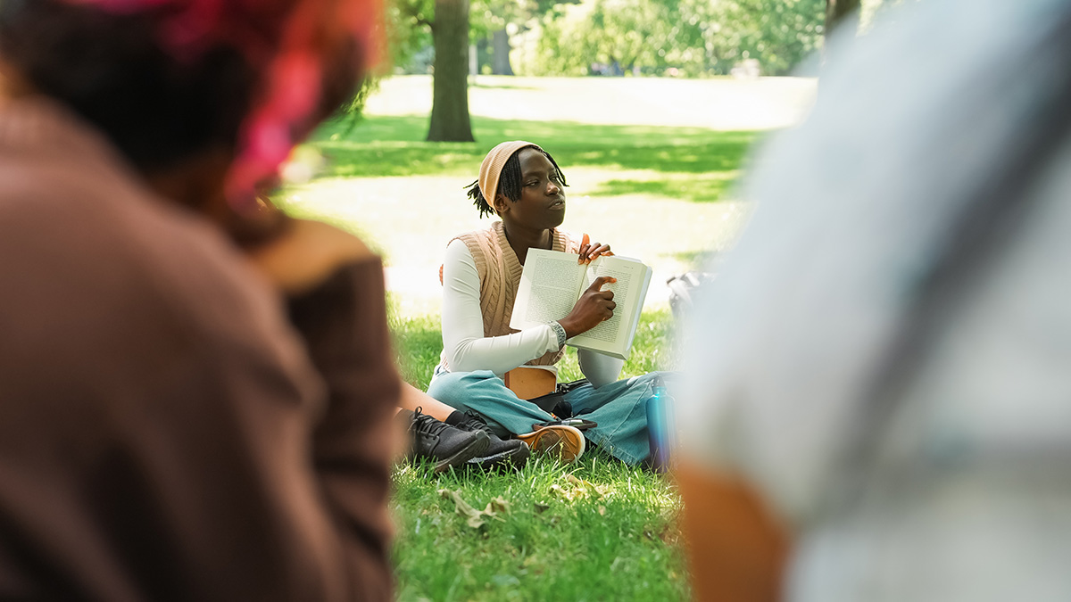 A person sitting cross-legged in a grassy park pointing to a passage in an open book.