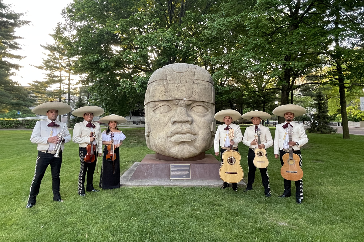 A mariachi band posing around giant stone Olmec head sculpture outdoors at Lehman College