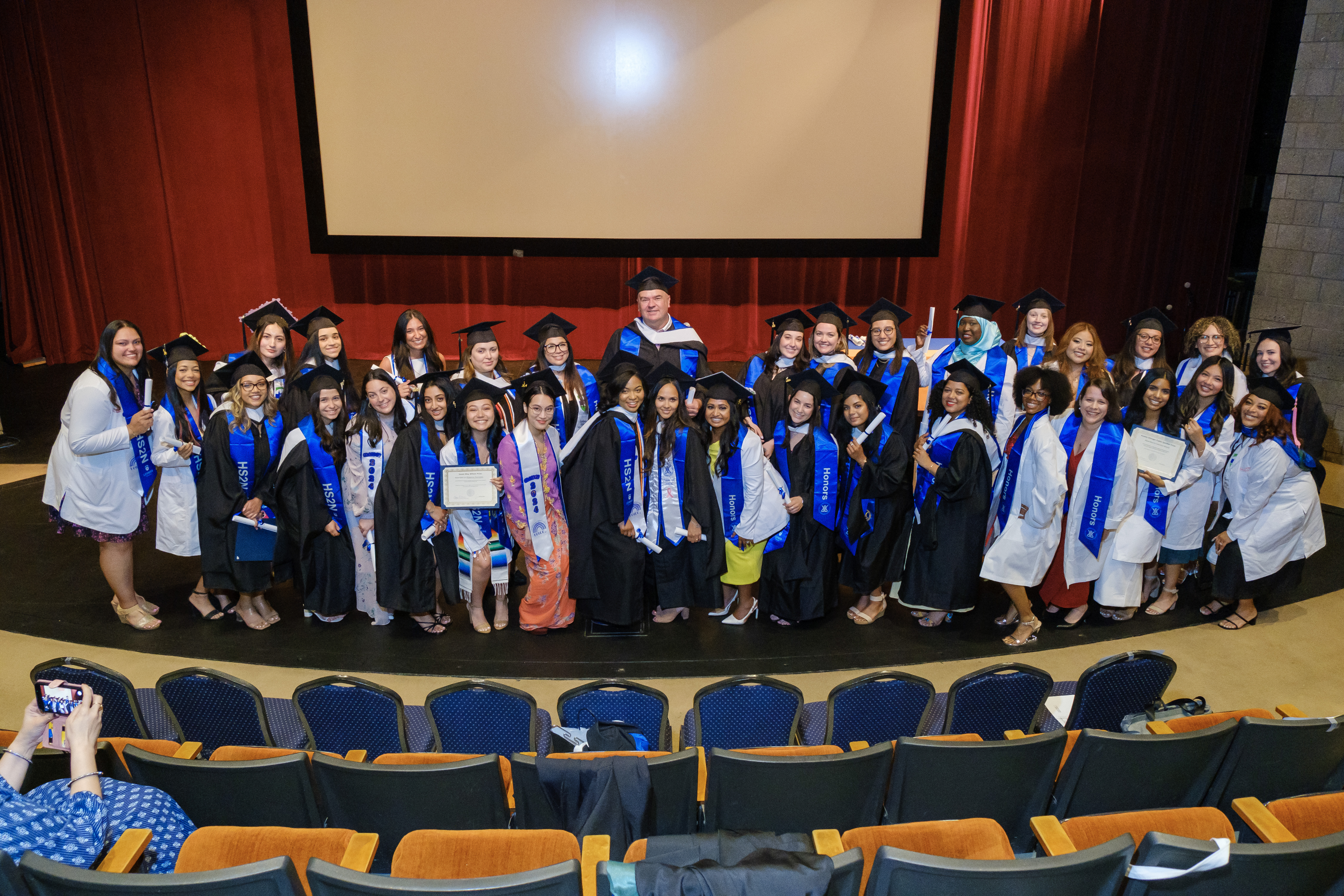A group of Speech graduates in their white coats, posing on a stage.